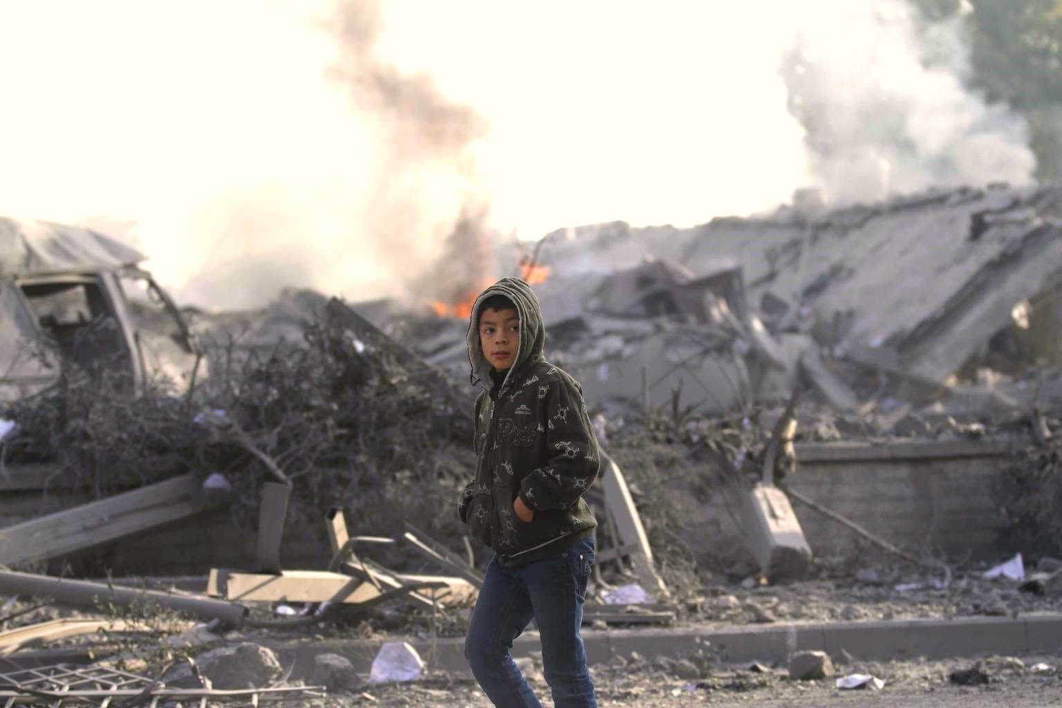 Un niño camina por su barrio en Gaza el cual se observa completamente destruido. Se observa fuego al fondo de la imagen